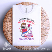 Футболка скільки би не стукнуло, білого кольору, зображена зверху на плетеній кошику, фон світлий, принт з персонажем у боксерських рукавичках, який б'є по тортові, текст на українській мові.