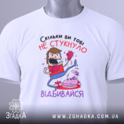 Футболка скільки би не стукнуло, білого кольору, зображена зверху, на ній видно етикетку, фон світло-сірий, принт з персонажем у боксерських рукавичках, який б'є по тортові, текст на українській мові.