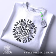 Футболка Слава Україні, білого кольору, складена на поверхні, фон світло-сірий, принт чорного кольору з тризубом та написом 'Слава Україні! Героям Слава!', на футболці є етикетка з зеленим шнурком.