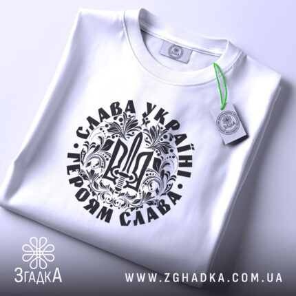 Футболка Слава Україні, білого кольору, складена на поверхні, фон світло-сірий, принт чорного кольору з тризубом та написом 'Слава Україні! Героям Слава!', на футболці є етикетка з зеленим шнурком.