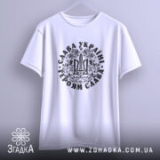 Футболка Слава Україні, білого кольору, підвішена на вішалці, фон світло-сірий, принт чорного кольору з тризубом та написом 'Слава Україні! Героям Слава!', рукави футболки короткі.