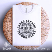 Футболка Слава Україні, білого кольору, складена на плетеній кошику, фон білий, принт чорного кольору з тризубом та написом 'Слава Україні! Героям Слава!', видна етикетка з написом.