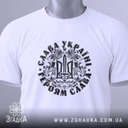 Футболка Слава Україні, білого кольору, складена на поверхні, фон світло-сірий, принт чорного кольору з тризубом та написом 'Слава Україні! Героям Слава!', на футболці є етикетка з зеленим шнурком.