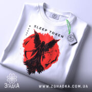 Футболка Sleep Token білого кольору, складена на поверхні, з принтом червоного кольору, що зображає фігуру з крилами на фоні червоного кола, видно етикетку з зеленим шнурком, фон світло-сірий.