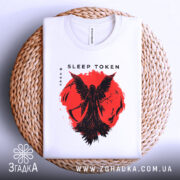 Футболка Sleep Token білого кольору, складена на плетеній кошику, з принтом червоного кольору, що зображає фігуру з крилами на фоні червоного кола, фон світло-сірий.