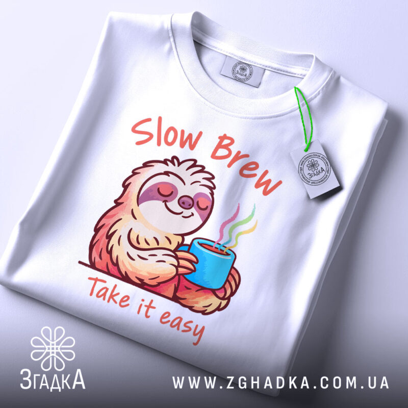 Футболка Slow Brew релакс білого кольору, зображена зверху, на ній видно принт з ведмедем-лінивцем, який тримає чашку з парою, текст 'Slow Brew' і 'Take it easy' яскраво виділяється, на футболці є етикетка з зеленим шнурком.