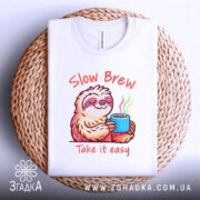 Футболка Slow Brew релакс білого кольору, зображена зверху на плетеній основі, фон світлий, принт з ведмедем-лінивцем, який тримає чашку з парою, текст 'Slow Brew' і 'Take it easy' яскраво виділяється на фоні.