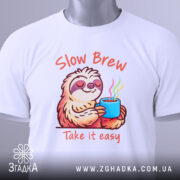 Футболка Slow Brew релакс білого кольору, зображена зверху, на ній видно принт з ведмедем-лінивцем, який тримає чашку з парою, текст 'Slow Brew' і 'Take it easy' яскраво виділяється, на футболці є етикетка з зеленим шнурком.