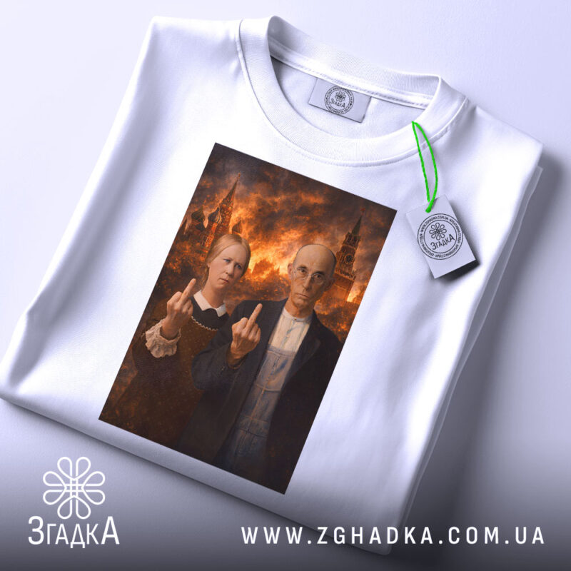 Футболка сміливий політичний принт, біла, зображена на столі, фон світлий, принт з двома персонажами, які показують середні пальці, прикріплена бірка з зеленим шнурком.