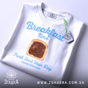 Футболка сніданок новий день, біла, складена на поверхні, фон світло-сірий, принт з написом 'Breakfast Time' та зображенням хліба з шоколадом, з етикеткою.