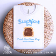Футболка сніданок новий день, біла, складена на кошику з плетеного матеріалу, фон світло-сірий, принт з написом 'Breakfast Time' та зображенням хліба з шоколадом.