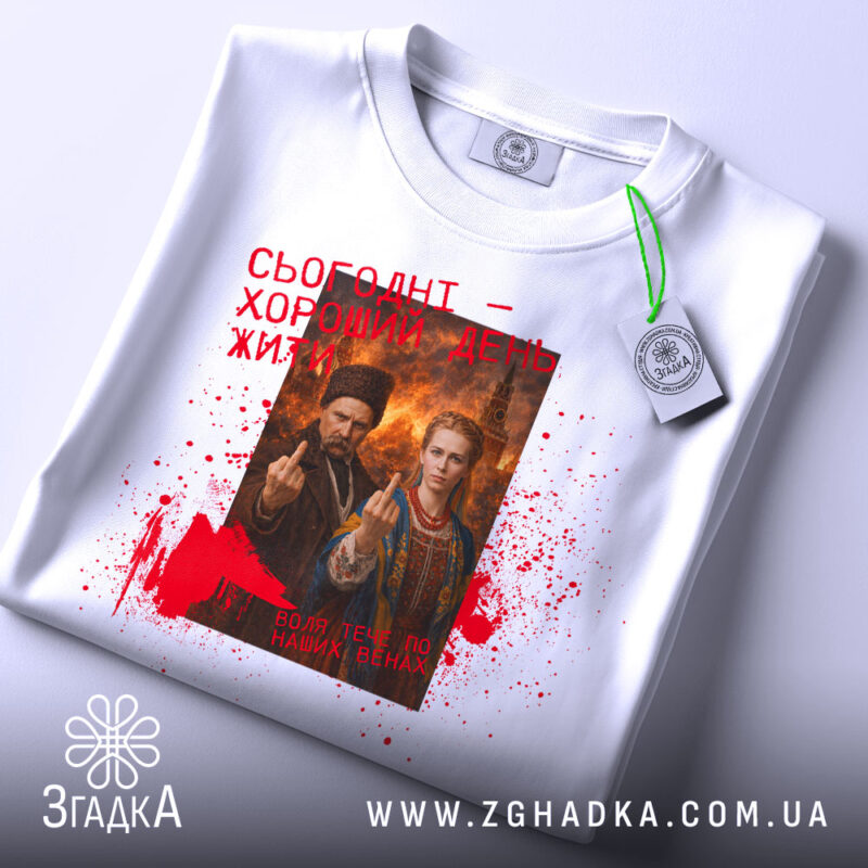 Футболка сьогодні хороший день. Біла футболка, розкладена на поверхні, з видимим етикеткою. Принт на футболці той же, що й на попередній, з червоними плямами та текстом. Фон нейтральний, що підкреслює дизайн футболки.