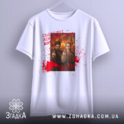 Футболка сьогодні хороший день. Біла футболка, підвішена на вішалці, з фронтального ракурсу. Принт з двома персонажами та червоними плямами, текст на футболці також червоний. Фон однорідний, що акцентує увагу на футболці.