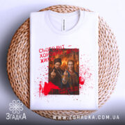 Футболка сьогодні хороший день. Біла футболка, складена на плетеній основі, з видимим принтом. Дизайн футболки повторює попередні, з червоними плямами та текстом. Фон натуральний, що створює затишну атмосферу.