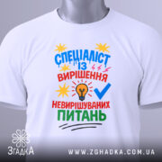 Футболка спеціаліст із питань білого кольору, зображена зверху, на плоскій поверхні, фон світло-сірий, принт яскравий з текстом та графічними елементами, на комірці видно етикетку.