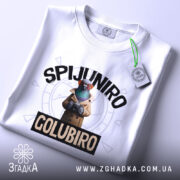 Футболка Spijuniro Golubiro білого кольору, зображена зверху, на столі, фон світло-сірий, принт з пташкою в окулярах та плащі, текст 'SPIJUNIRO' зверху та 'GOLUBIRO' внизу, на футболці є етикетка.