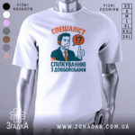 Футболка спілкування з довбойобами, біла, зображена на манекені, фон світло-сірий, принт з чоловіком у навушниках, який піднімає великий палець, текст червоного та зеленого кольорів.