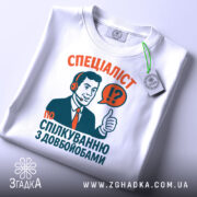 Футболка спілкування з довбойобами, біла, зображена зверху, фон світло-сірий, принт з чоловіком у навушниках, який піднімає великий палець, текст червоного та зеленого кольорів, на футболці є етикетка.