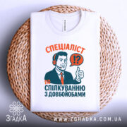 Футболка спілкування з довбойобами, біла, зображена на плетеній кошику, фон світло-сірий, принт з чоловіком у навушниках, який піднімає великий палець, текст червоного та зеленого кольорів.