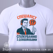 Футболка спілкування з довбойобами, біла, зображена зверху, фон світло-сірий, принт з чоловіком у навушниках, який піднімає великий палець, текст червоного та зеленого кольорів, на футболці є етикетка.