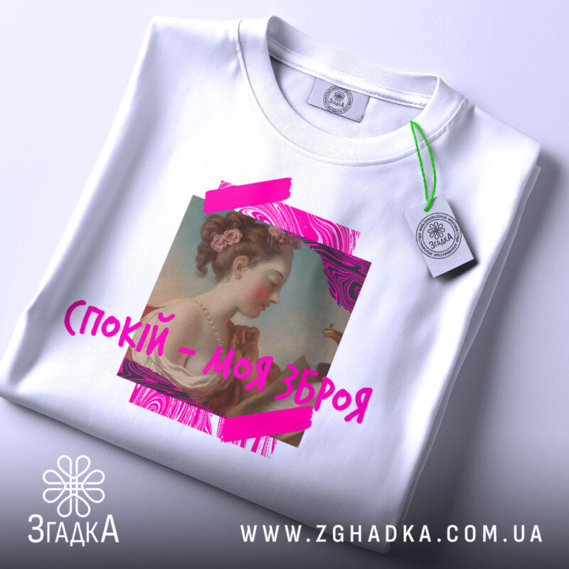 Футболка спокій моя зброя, біла, складена на поверхні, фон світлий, принт з портретом жінки в профіль, яскраві рожеві елементи навколо.