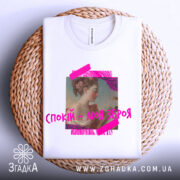 Футболка спокій моя зброя, біла, складена на плетеній кошику, фон світлий, принт з портретом жінки в профіль, яскраві рожеві елементи навколо.