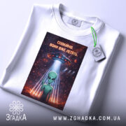 Футболка спокійно вони летять, біла, складена на поверхні, з видимим етикеткою, фон світлий, принт з зеленим інопланетянином, який показує знак миру, під космічним кораблем, оточеним зірками та планетами.