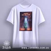 Футболка спокійно вони летять, біла, підвішена на вішалці, фон світлий, принт з зеленим інопланетянином, який показує знак миру, під космічним кораблем, оточеним зірками та планетами.