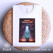 Футболка спокійно вони летять, біла, складена на плетеній кошику, фон світлий, принт з зеленим інопланетянином, який показує знак миру, під космічним кораблем, оточеним зірками та планетами.