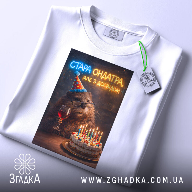 Футболка стара ондатра, біла, зображена з верхнього ракурсу, на фоні світлого градієнта, принт з ведмедем у святковому капелюшку, що тримає келих з вином, поряд з тортом з свічками, на комірці є етикетка.