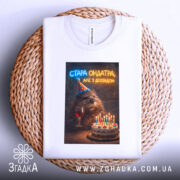 Футболка стара ондатра, біла, зображена зверху на плетеній основі, фон світлий, принт з ведмедем у святковому капелюшку, що тримає келих з вином, поряд з тортом з свічками.