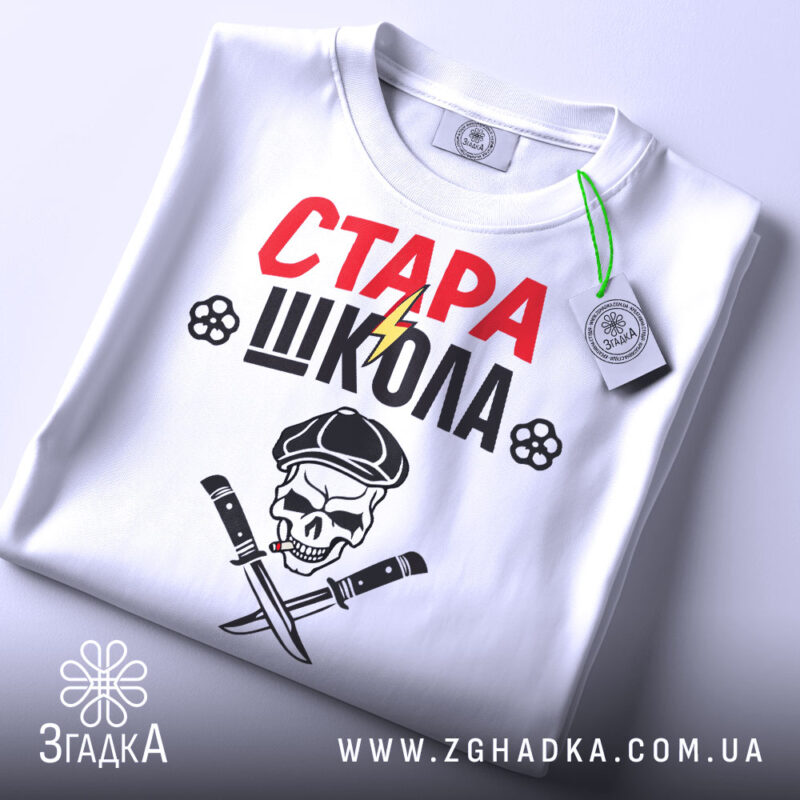 Футболка стара школа, біла, складена на поверхні, фон світлий, принт з червоним написом 'СТАРА' та чорним 'ШКОЛА', зображення черепа з капелюхом та ножами, на футболці є етикетка.