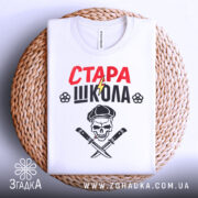 Футболка стара школа, біла, складена на плетеній основі, фон світлий, принт містить червоний напис 'СТАРА' та чорний 'ШКОЛА', зображення черепа з капелюхом та ножами, футболка акуратно розкладена.