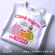 Футболка старій ондатрі білого кольору, складена на поверхні, фон світло-сірий. Принт з малюнками пряжі, чашки з напоєм та спиць, текст 'З Днем народження від подруги' яскравий, з елементами декору.