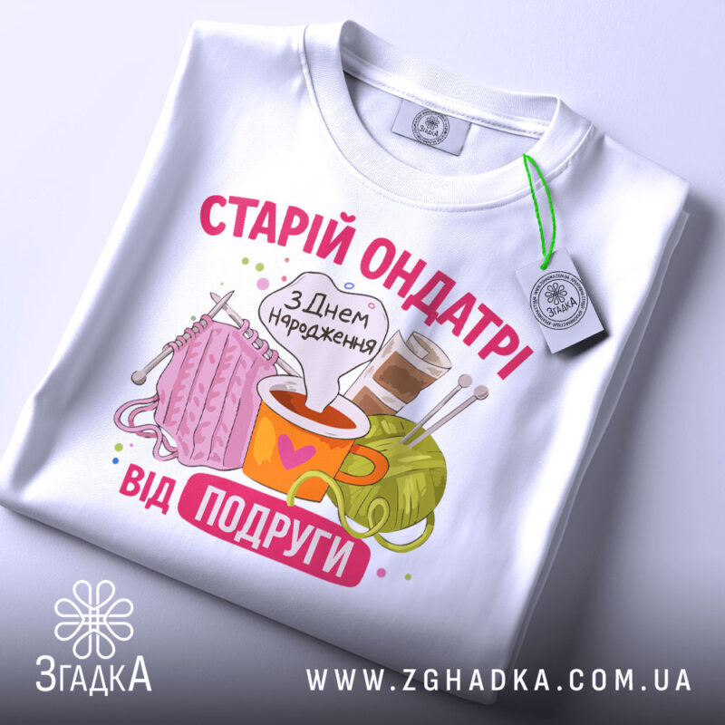 Футболка старій ондатрі білого кольору, складена на поверхні, фон світло-сірий. Принт з малюнками пряжі, чашки з напоєм та спиць, текст 'З Днем народження від подруги' яскравий, з елементами декору.