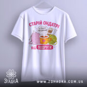 Футболка старій ондатрі білого кольору, зображена з фронтального ракурсу на манекені, фон світло-сірий. Принт містить малюнки пряжі, чашки з напоєм та спиць, текст 'З Днем народження від подруги' у яскравих кольорах.