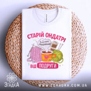 Футболка старій ондатрі білого кольору, складена на плетеній кошику, фон світло-сірий. Принт містить малюнки пряжі, чашки з напоєм та спиць, текст 'З Днем народження від подруги' у яскравих кольорах.