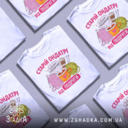 Футболка старій ондатрі білого кольору, зображена з фронтального ракурсу на манекені, фон світло-сірий. Принт містить малюнки пряжі, чашки з напоєм та спиць, текст 'З Днем народження від подруги' у яскравих кольорах.