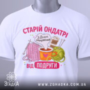 Футболка старій ондатрі білого кольору, складена на поверхні, фон світло-сірий. Принт з малюнками пряжі, чашки з напоєм та спиць, текст 'З Днем народження від подруги' яскравий, з елементами декору.