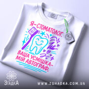 Футболка стоматолог білого кольору, складена на поверхні, фон світлий, принт з текстом "Я - стоматолог. Ваша усмішка - мій автограф" у яскравих кольорах, з малюнком зуба та зубної щітки, на етикетці присутній логотип.