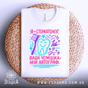 Футболка стоматолог білого кольору, складена на плетеній основі, фон світлий, принт містить текст "Я - стоматолог. Ваша усмішка - мій автограф" у яскравих кольорах, з малюнком зуба та зубної щітки, виглядає естетично.