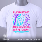 Футболка стоматолог білого кольору, складена на поверхні, фон світлий, принт з текстом "Я - стоматолог. Ваша усмішка - мій автограф" у яскравих кольорах, з малюнком зуба та зубної щітки, на етикетці присутній логотип.