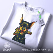 Футболка стоп війна, біла, зображена зверху, фон світлий, принт з доберманом, що тримає табличку 'STOP WAR', на футболці є етикетка з зеленим шнурком.