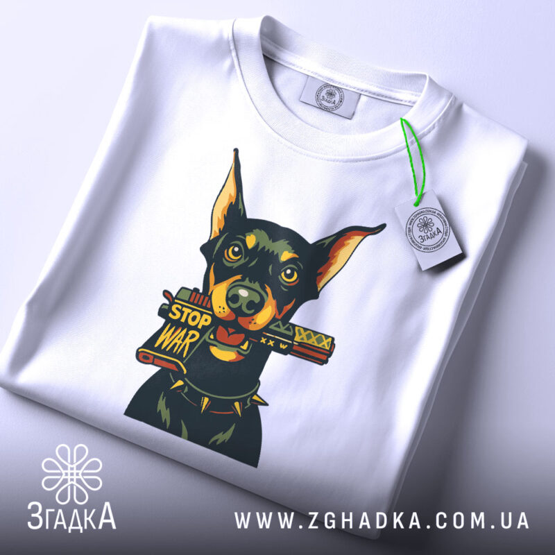 Футболка стоп війна, біла, зображена зверху, фон світлий, принт з доберманом, що тримає табличку 'STOP WAR', на футболці є етикетка з зеленим шнурком.