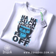 Футболка Such Sarcasm білого кольору, складена на столі, фон світло-сірий, принт з зображенням горили та текстом "HA HA SUCH SARCASM F*CK OFF" у чорному та блакитному кольорах, з біркою на зеленому шнурку.