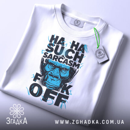 Футболка Such Sarcasm білого кольору, складена на столі, фон світло-сірий, принт з зображенням горили та текстом "HA HA SUCH SARCASM F*CK OFF" у чорному та блакитному кольорах, з біркою на зеленому шнурку.