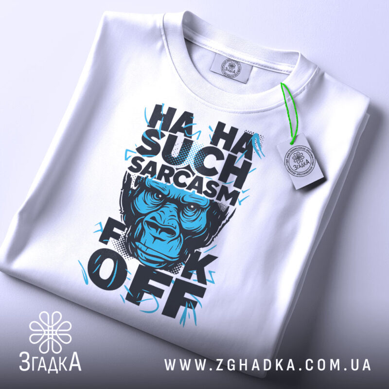 Футболка Such Sarcasm білого кольору, складена на столі, фон світло-сірий, принт з зображенням горили та текстом "HA HA SUCH SARCASM F*CK OFF" у чорному та блакитному кольорах, з біркою на зеленому шнурку.