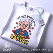 Футболка супер бабуся, біла, зображена на столі, фон світло-сірий, принт з бабусею в червоному плащі, з написом 'СУПЕР БАБУСЯ' та зеленим етикеткою.