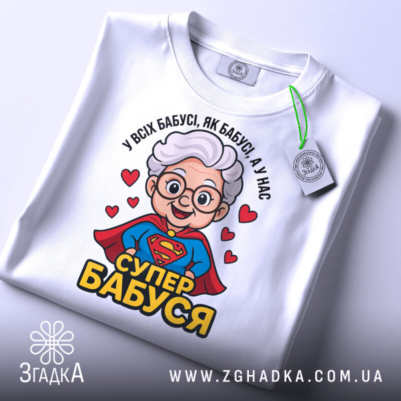 Футболка супер бабуся, біла, зображена на столі, фон світло-сірий, принт з бабусею в червоному плащі, з написом 'СУПЕР БАБУСЯ' та зеленим етикеткою.