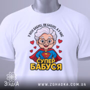 Футболка супер бабуся, біла, зображена на столі, фон світло-сірий, принт з бабусею в червоному плащі, з написом 'СУПЕР БАБУСЯ' та зеленим етикеткою.